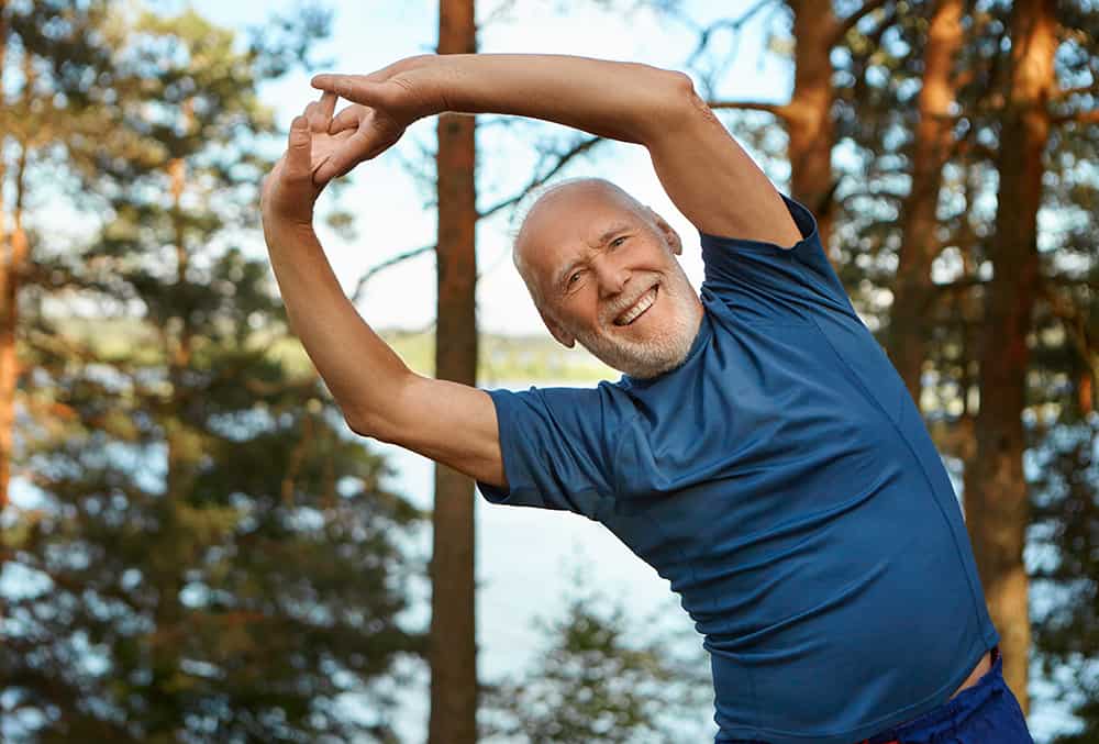 outdoor shot happy energetic senior retired man enjoying physical training park doing side bends exercise holding hands together with broad smile warming up body before run